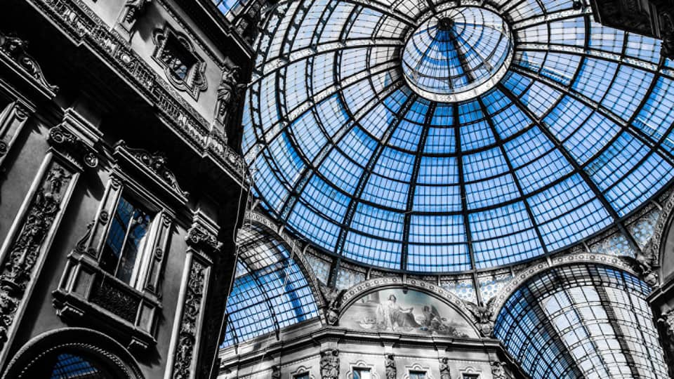 Interior view of the Galleria Vittorio Emanuele II in Milan, showcasing its grand glass dome ceiling and ornate architectural details in a mix of black and white with blue-tinted glass.