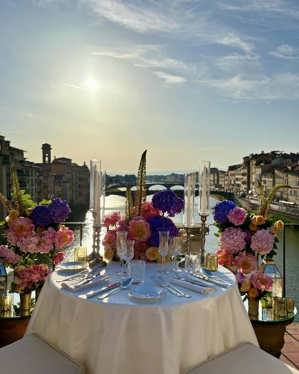 An elegantly set outdoor dining table overlooking a a river, with a sunny sky in the background.