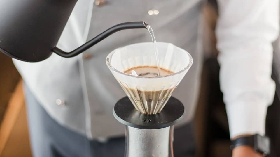 Barista holds black teapot, pours hot water over coffee in cone filter and glass jug on tray