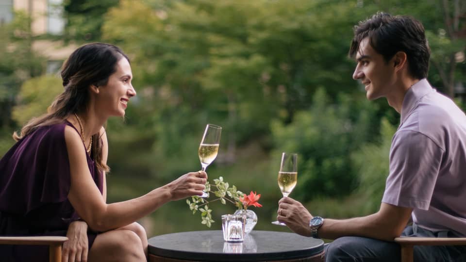 Two people sitting outdoors, toasting with champagne glasses at a small table with a flower vase