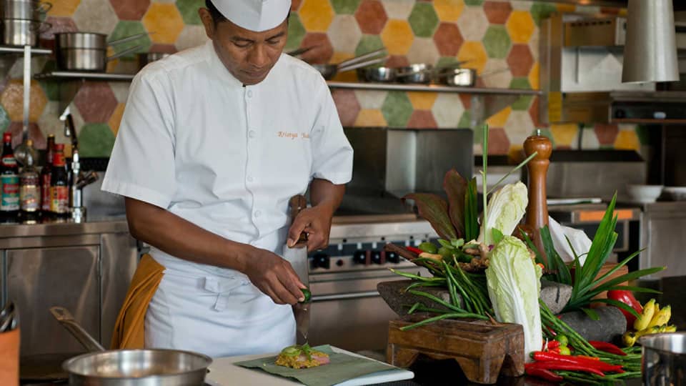 Chef in white uniform squeezes lime on large kitchen knife over dish, next to heads of fresh lettuce and whole peppers