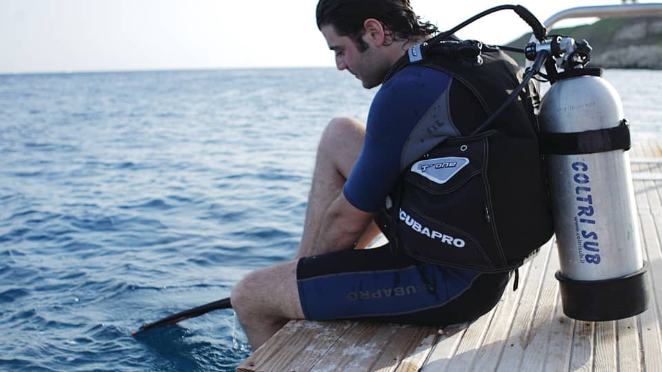 Man with wet hair wearing wetsuit, scuba diving tank sits on dock with feet in water 