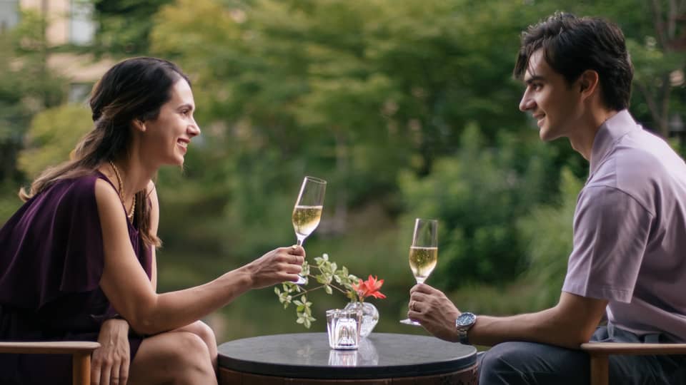 Two people sitting outdoors, toasting with champagne glasses at a small table with a flower vase