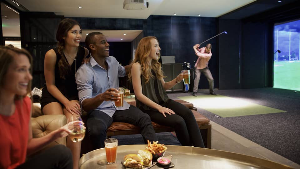 A man and three woman sip beer and cheer on a sports team in the foreground, while another man golfs in the background of a function room at four seasons hotel houston 