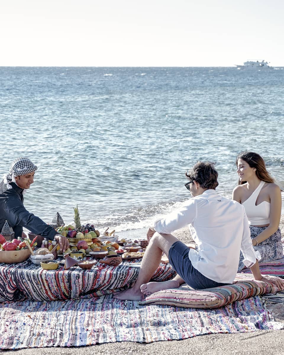 Three people sit on a beach around a low table set with a luxurious array of fruit, bread, and clay dishes full of food.