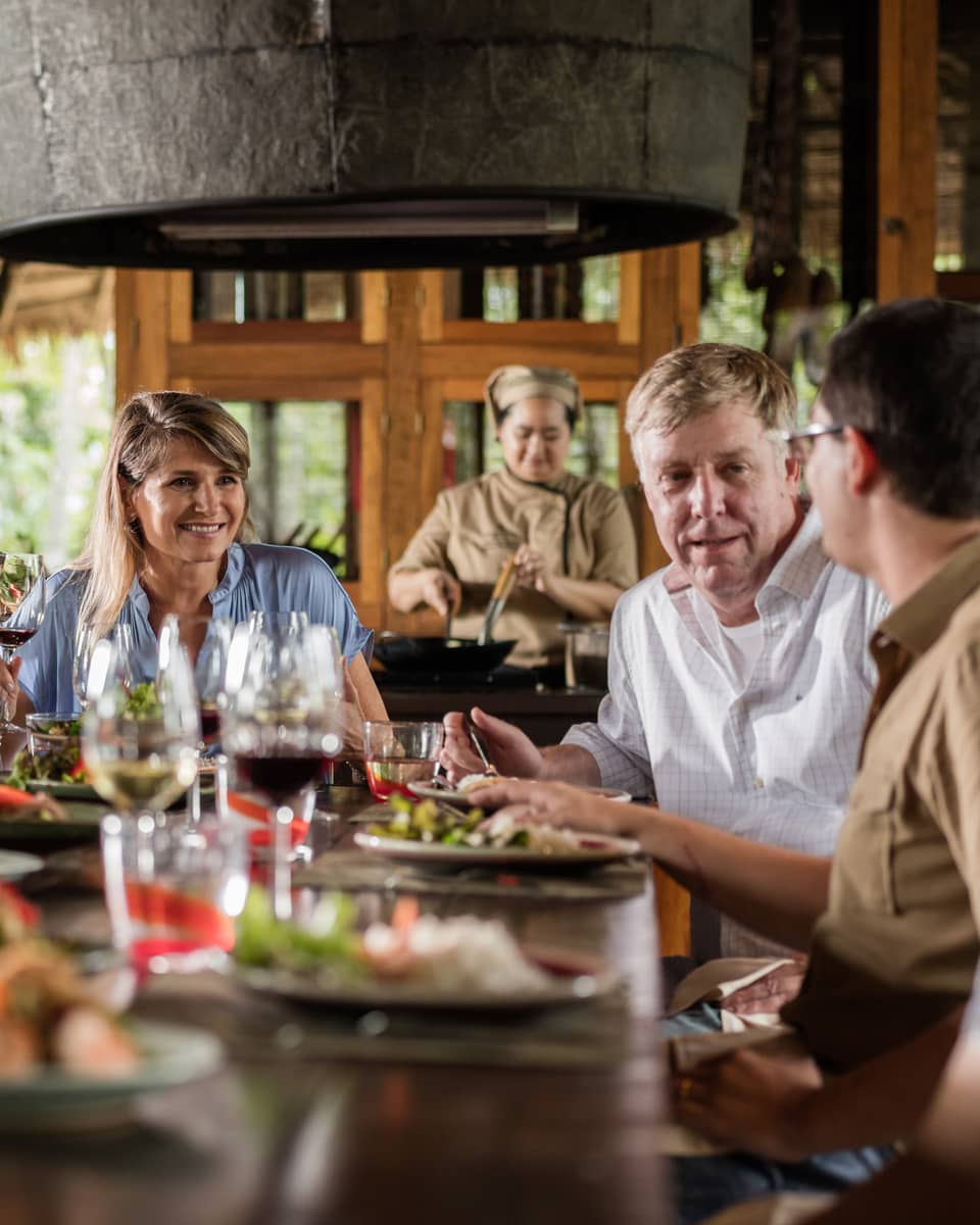A group around a table set with colourful fare and glasses of wine, as a chef in the background stirs a dish in a deep pan.