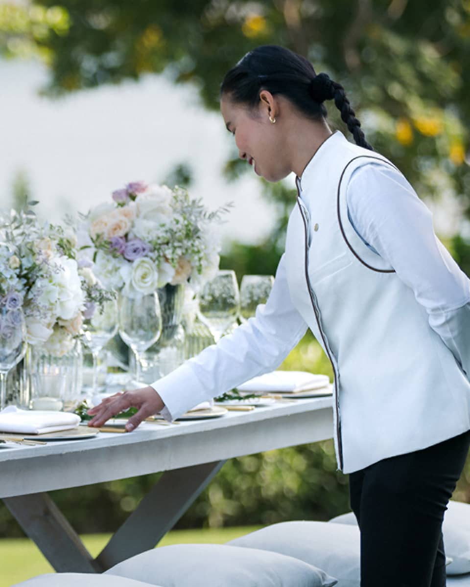A four seasons staff does the final touches on an outdoor, long banquet table decorated with large white floral arrangements 