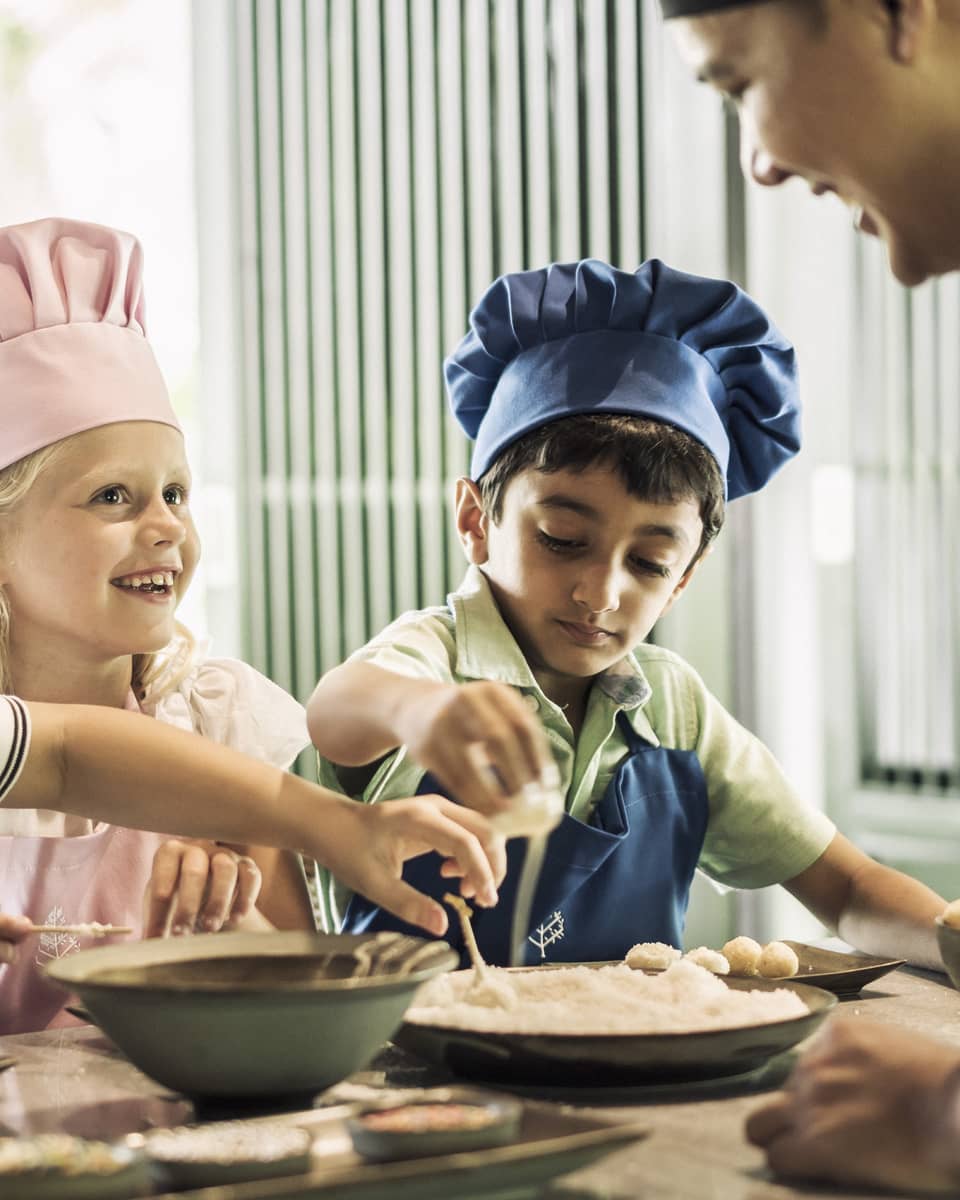 Three children with pink or blue chefs hats making food.