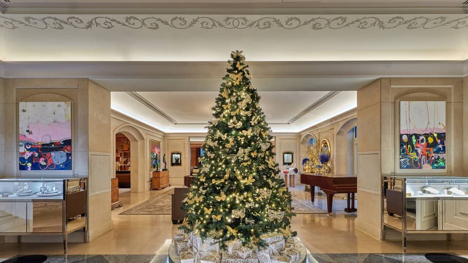 Luxurious hotel lobby, featuring a large elegantly decorated Christmas tree with gold ornaments and lights, surrounded by Christian Dior gift boxes and modern artwork on the walls.