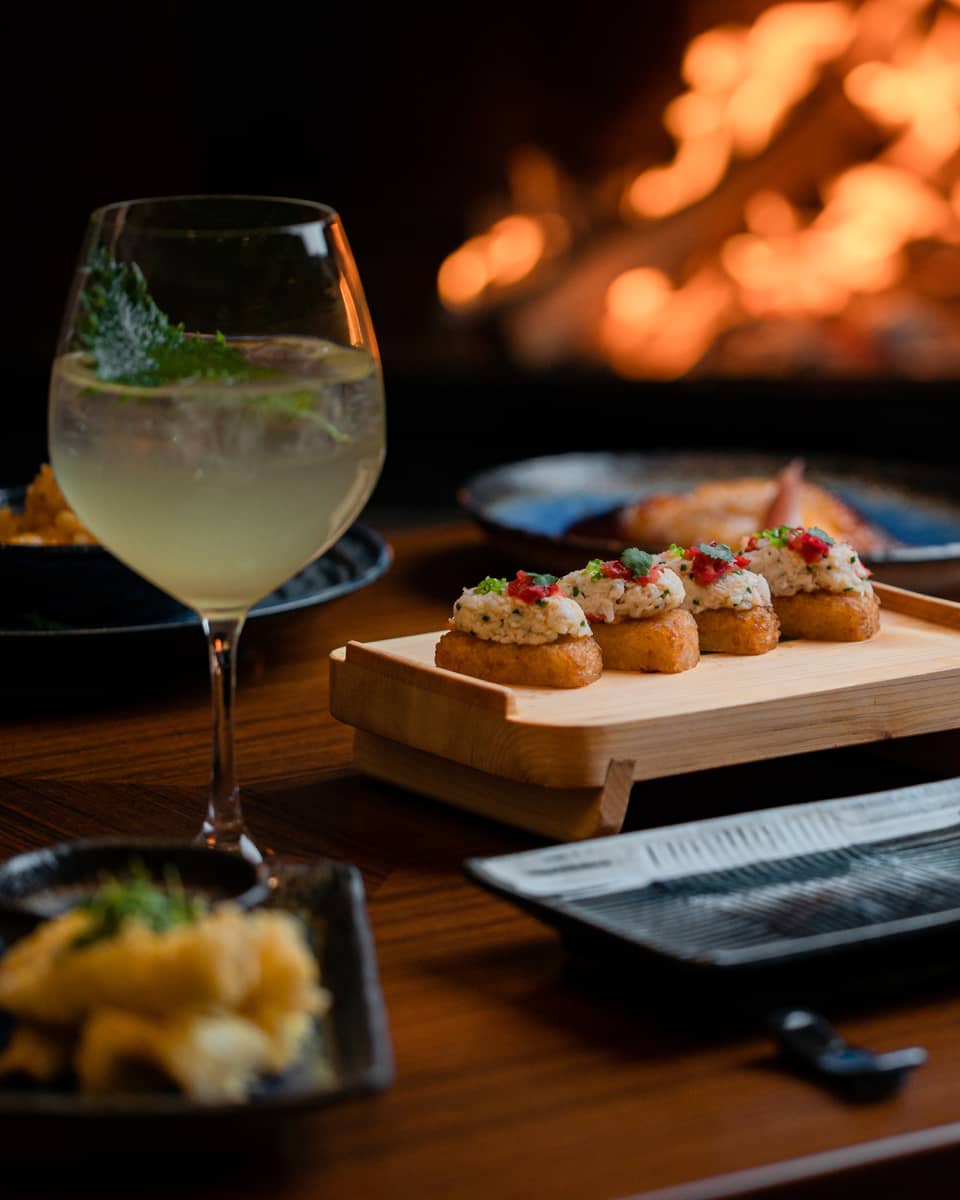 A cocktail and appetisers placed on a wooden table with a fireplace glowing in the background, creating a cosy dining atmosphere