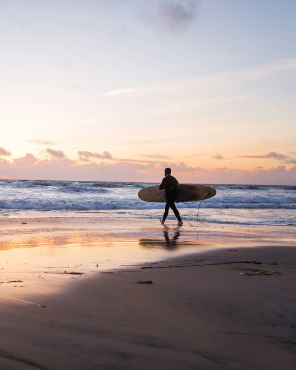 Under an expansive sky at sunset, clouds receding on the horizon, a surfer with a board heads into the glimmering ocean.