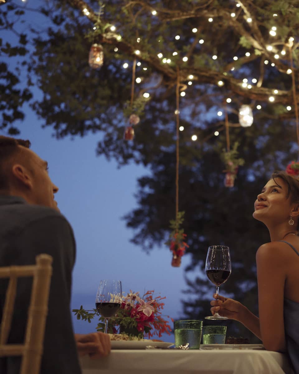 A smiling couple having a romantic dinner under trees covered in string lights and jars of flowers hanging from the branches.
