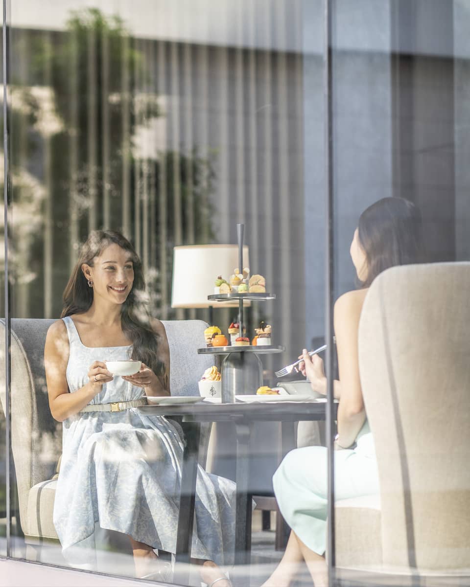 View through cafe glass window where two women enjoy afternoon tea and desserts