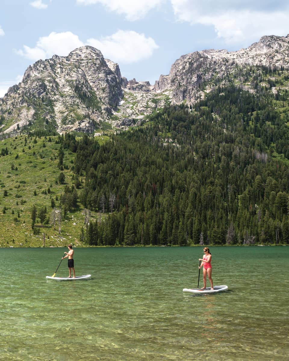 Two people on stand-up paddleboards on lake under mountain hills, trees, snowy peaks
