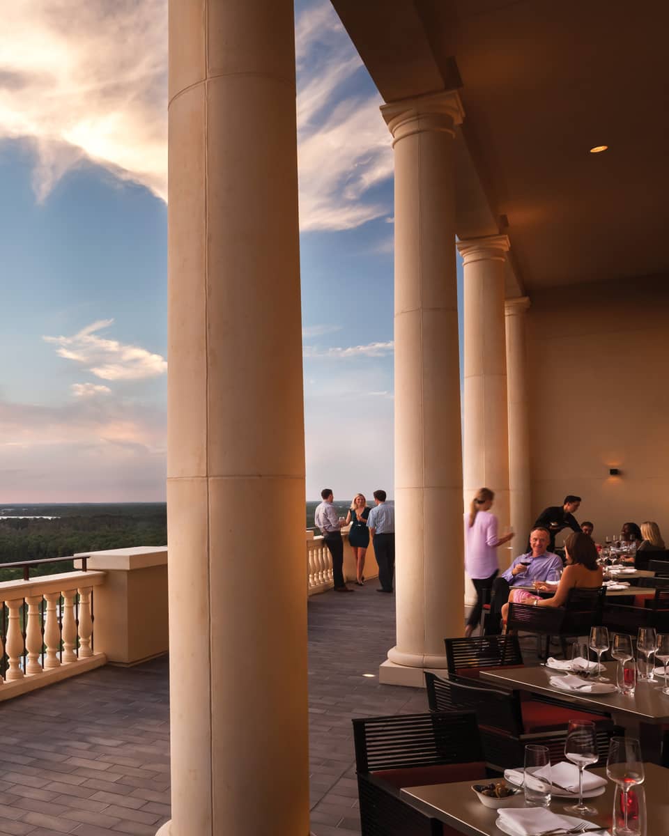 People dine at patio tables under tall pillars, balcony overlooking trees and sunset