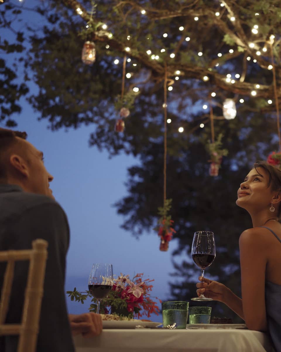 A smiling couple having a romantic dinner under trees covered in string lights and jars of flowers hanging from the branches.