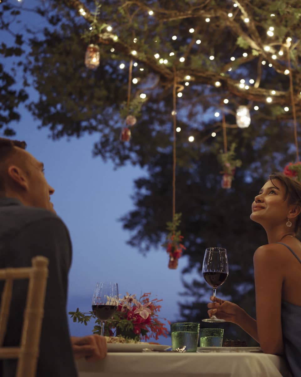 A smiling couple having a romantic dinner under trees covered in string lights and jars of flowers hanging from the branches.