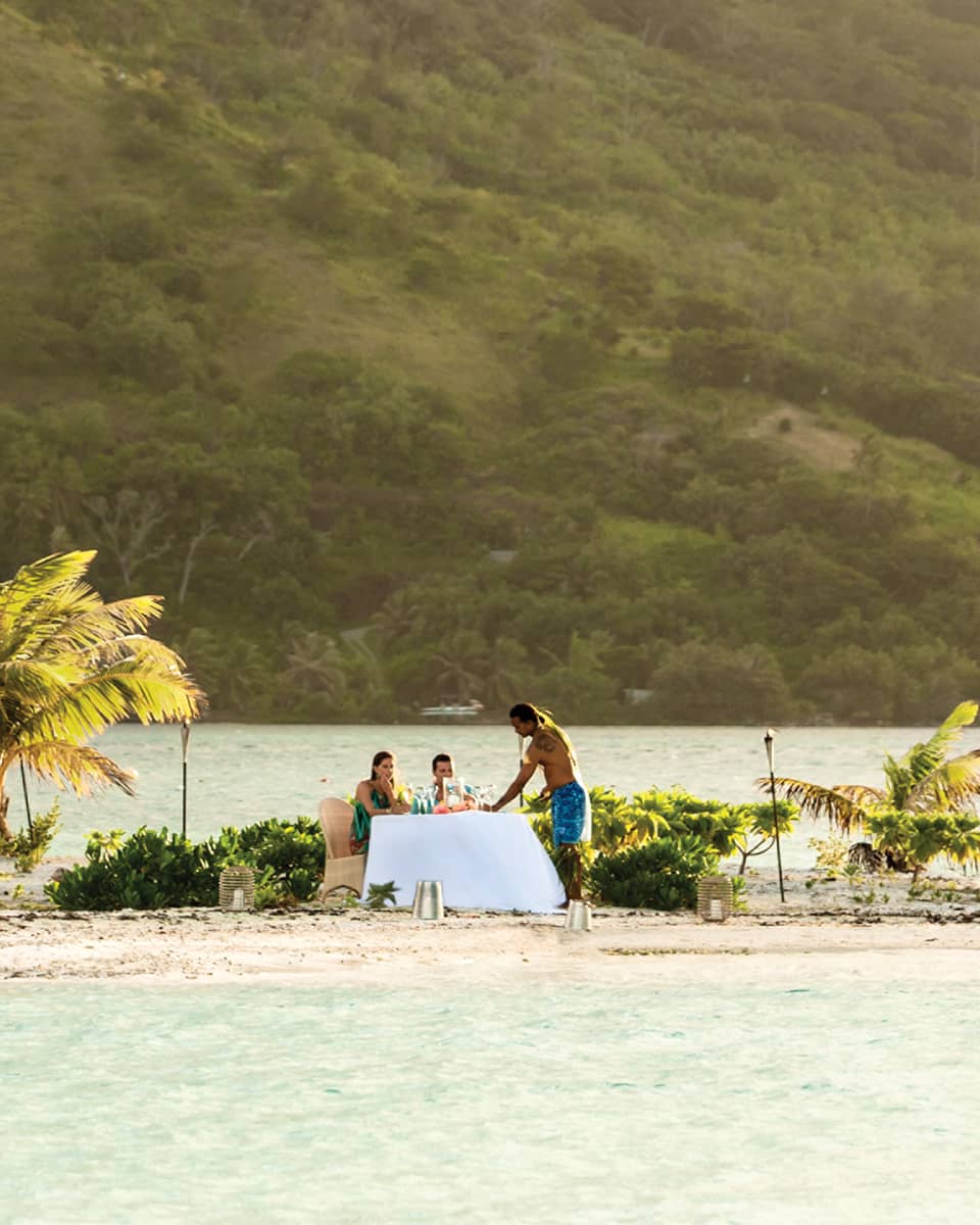 Small tropical island with palm trees, where two people are dining at a table by the water, with a mountain in the background