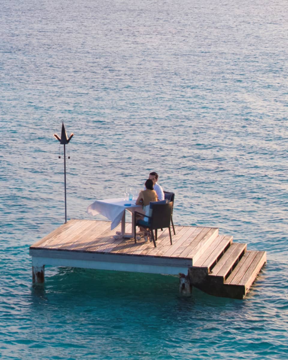 Man, woman dine at private table on small floating wood platform in middle of blue lagoon