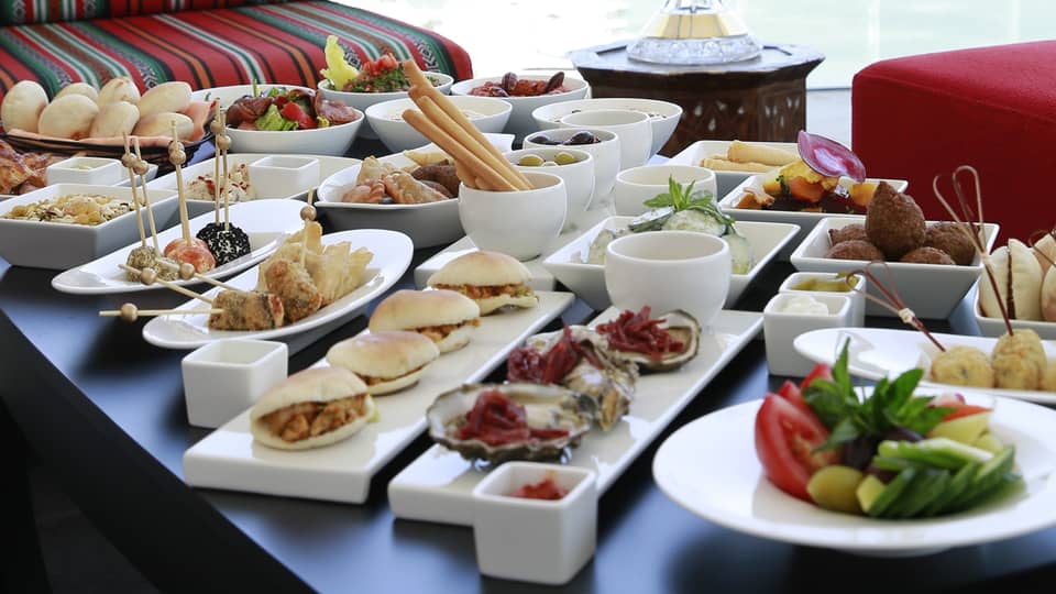 Table with more than a dozen white dishes with mezze appetizers and snacks, striped sofa and red bench