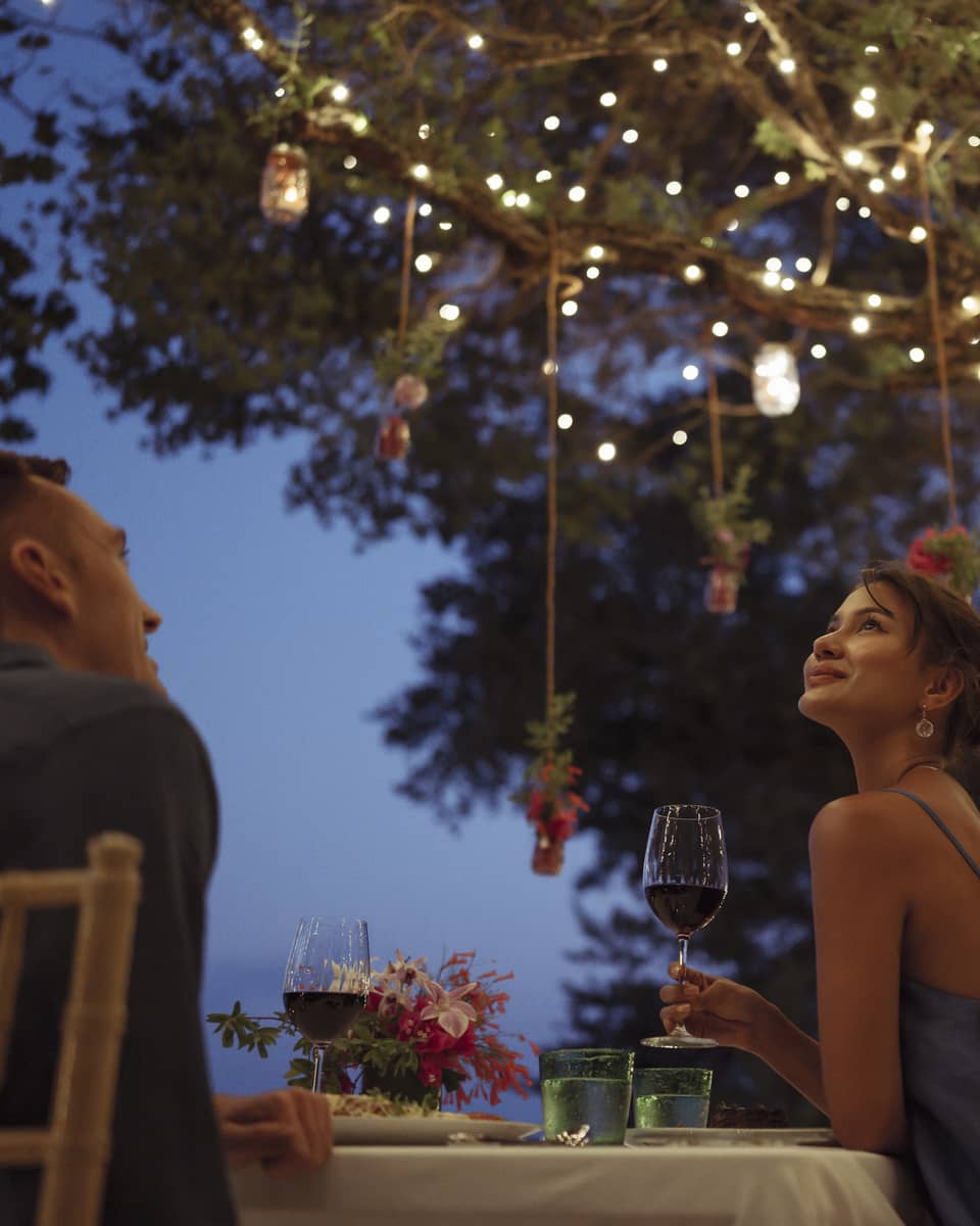 A smiling couple having a romantic dinner under trees covered in string lights and jars of flowers hanging from the branches.