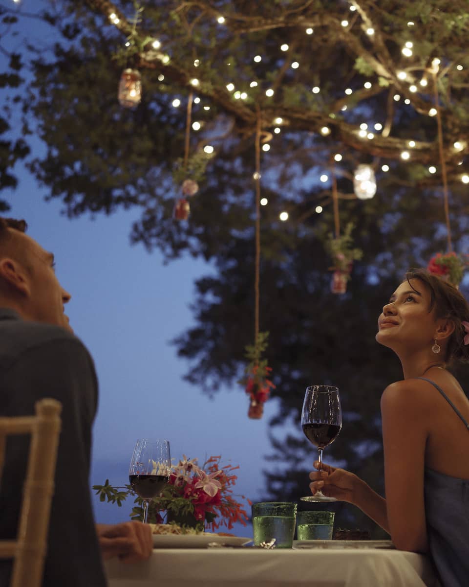 A smiling couple having a romantic dinner under trees covered in string lights and jars of flowers hanging from the branches.