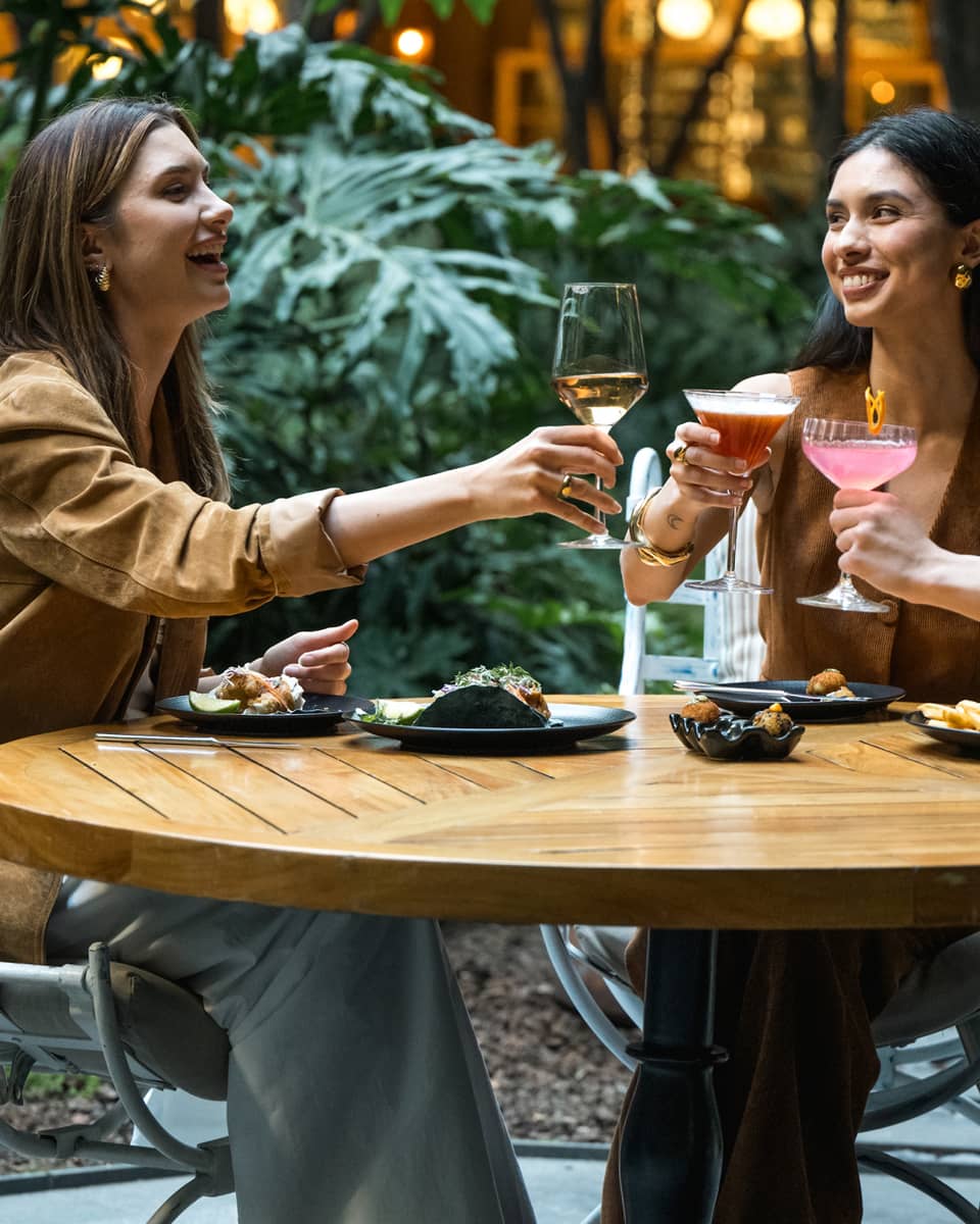 Three guests sit around a round wood table in an outdoor dining space and raise their glasses in a toast.