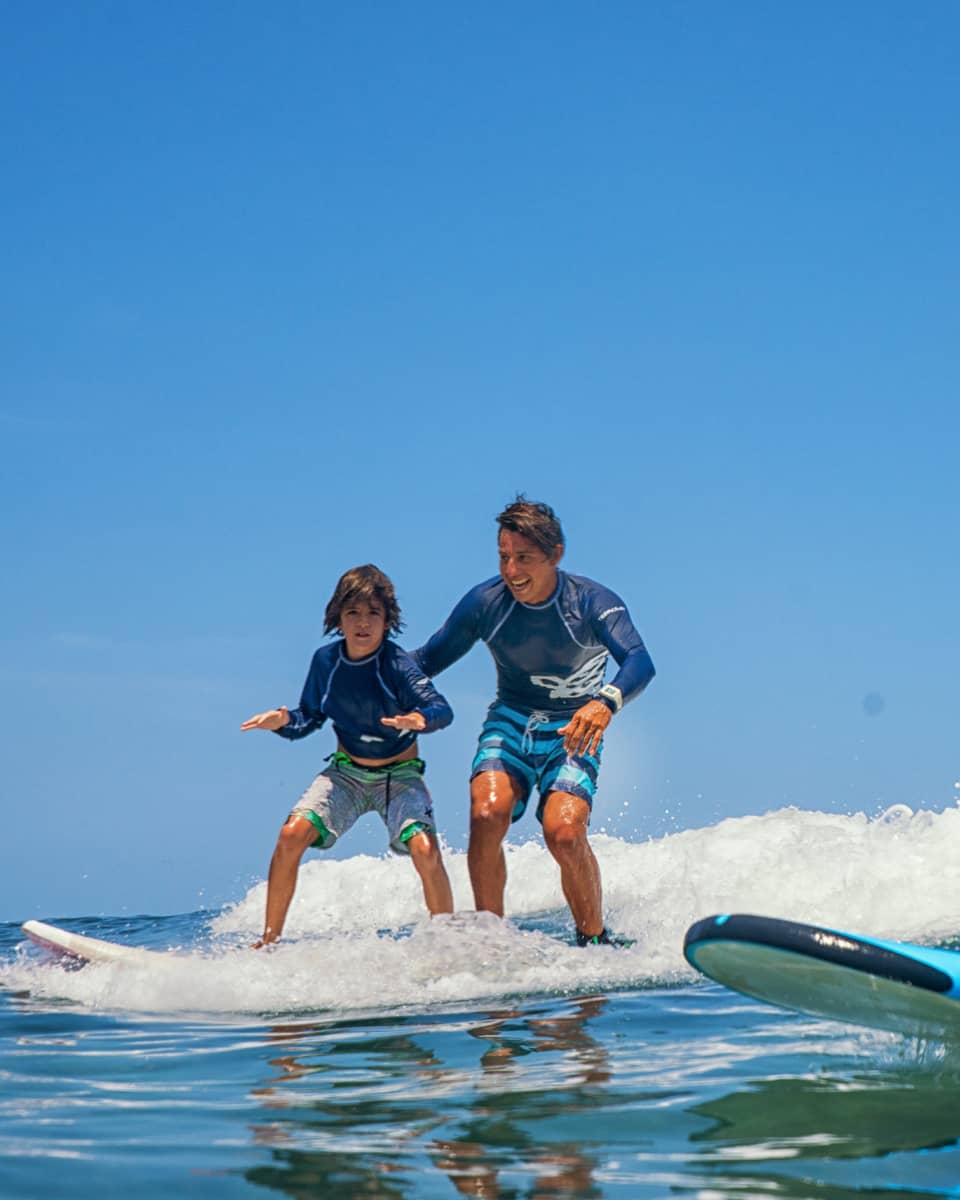 A pair of smiling adults and children standing on two surfboards, wearing swimsuits and rash guards, riding a small wave.