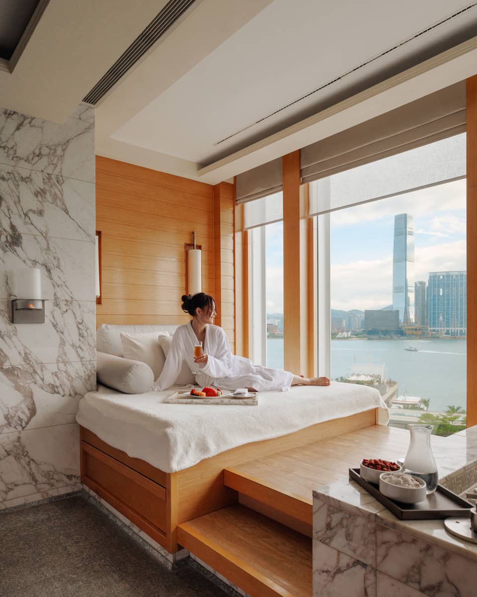 Woman in spa bathrobe sits on windowside chaise looking out to Victoria Harbour at Four Seasons Hotel Hong Kong