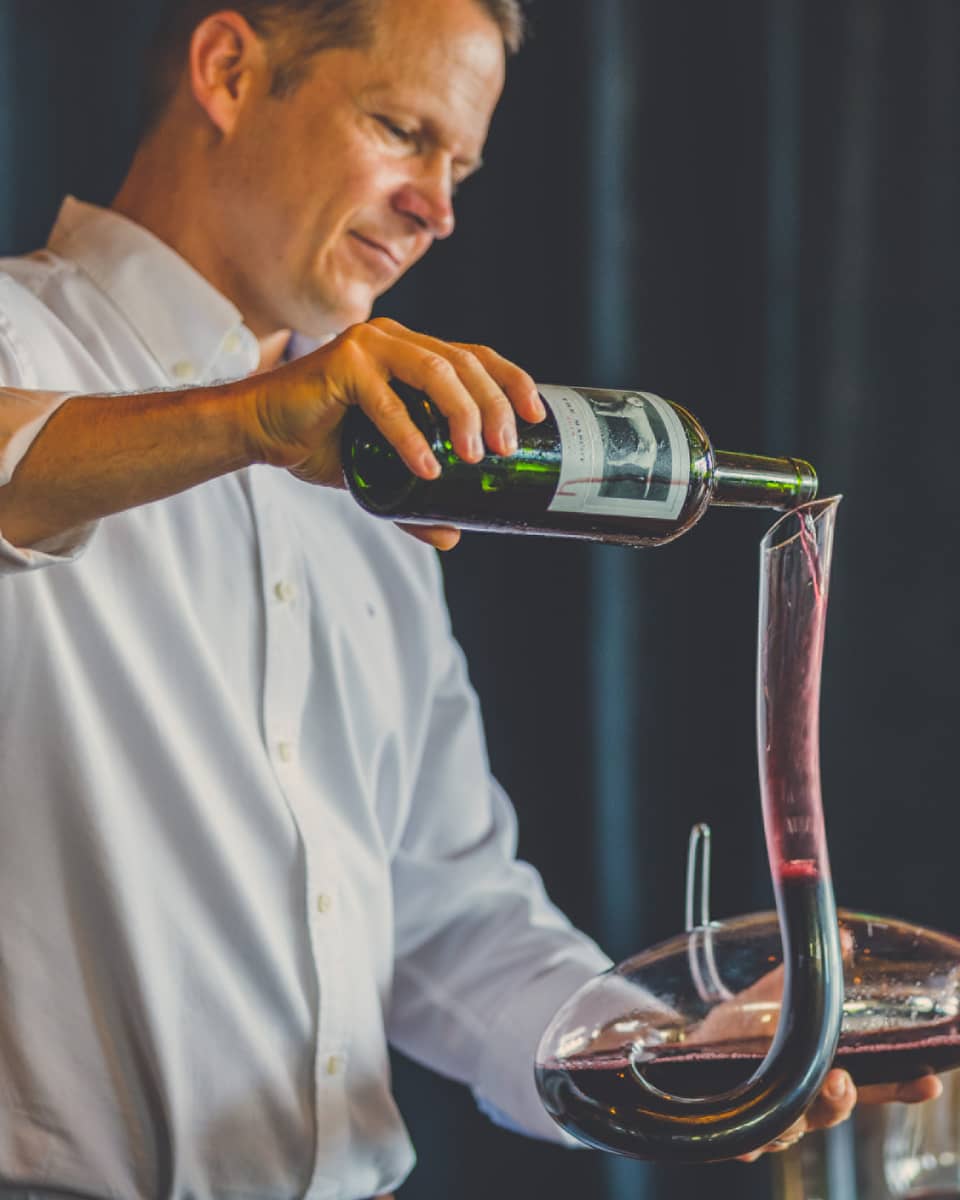 Man pours red wine into long, curved glass carafe near wine glasses