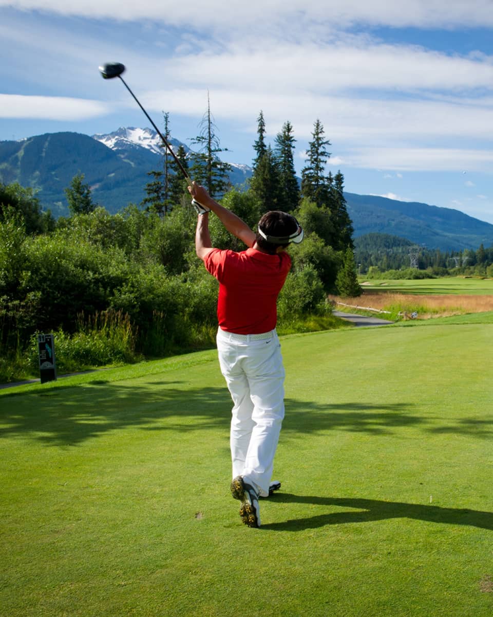 Back view of man swinging golf club on green, mountains in distance 