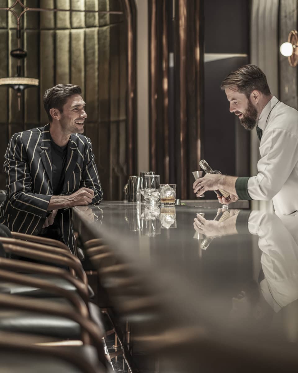 Bartender preparing a drink while a customer sits at the bar, both smiling, in a modern, upscale bar with a reflective countertop and warm lighting.
