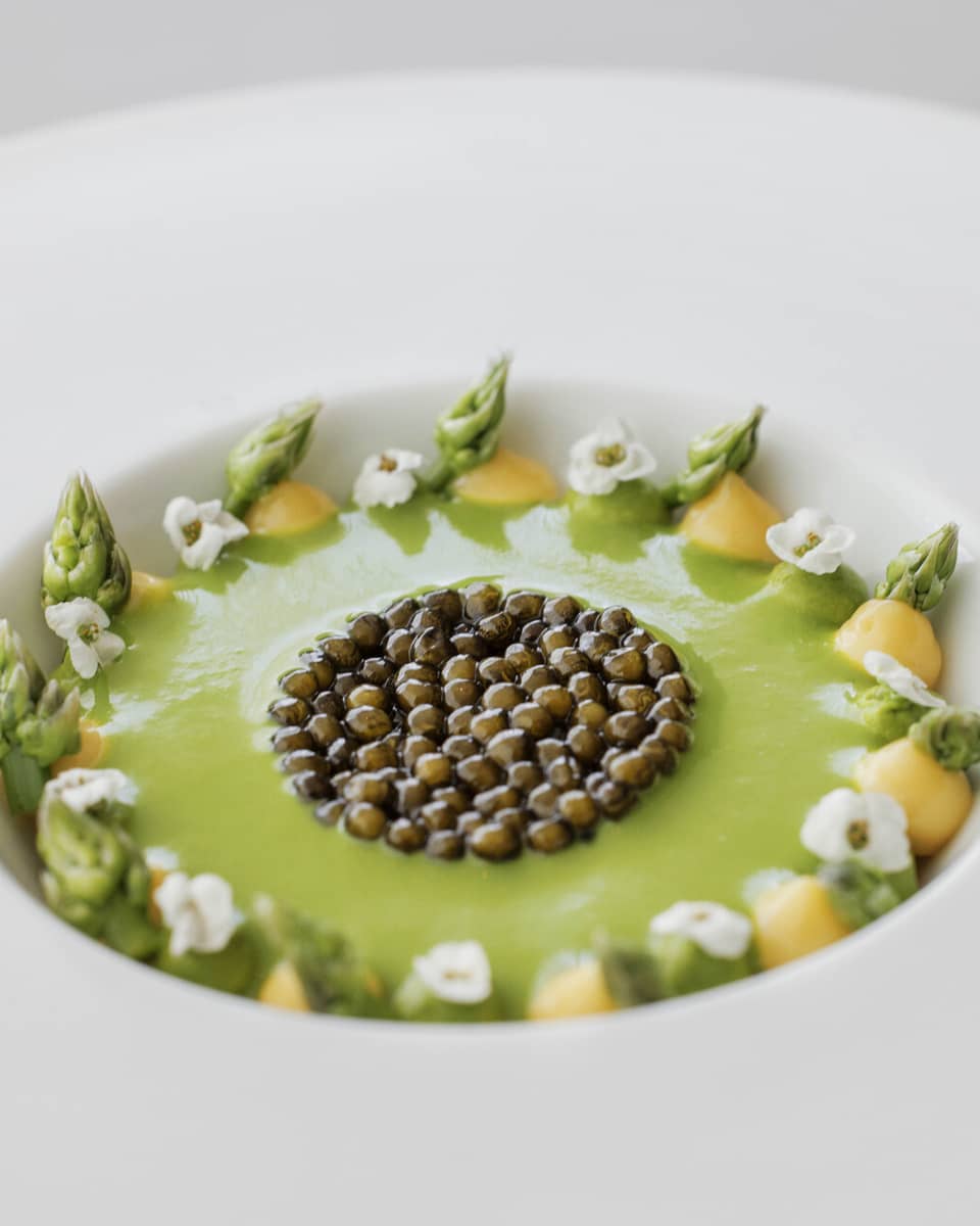 Caviar served over a green sauce in a bowl lined with dots of yellow sauce, asparagus and floral garnishes