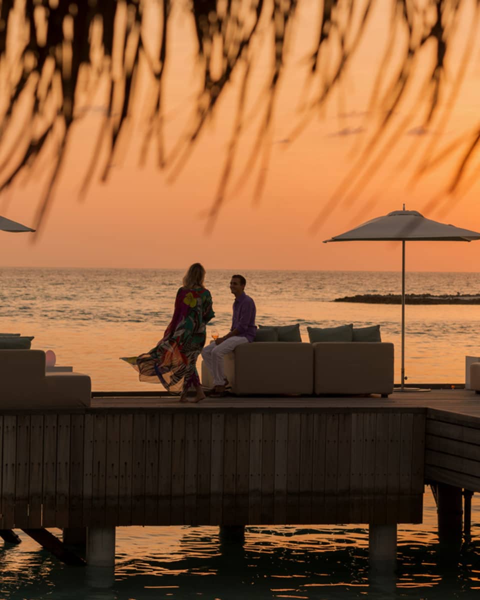 Private island dining at sunset. Woman with long dress blowing in the wind, man sits on the edge of white sofa