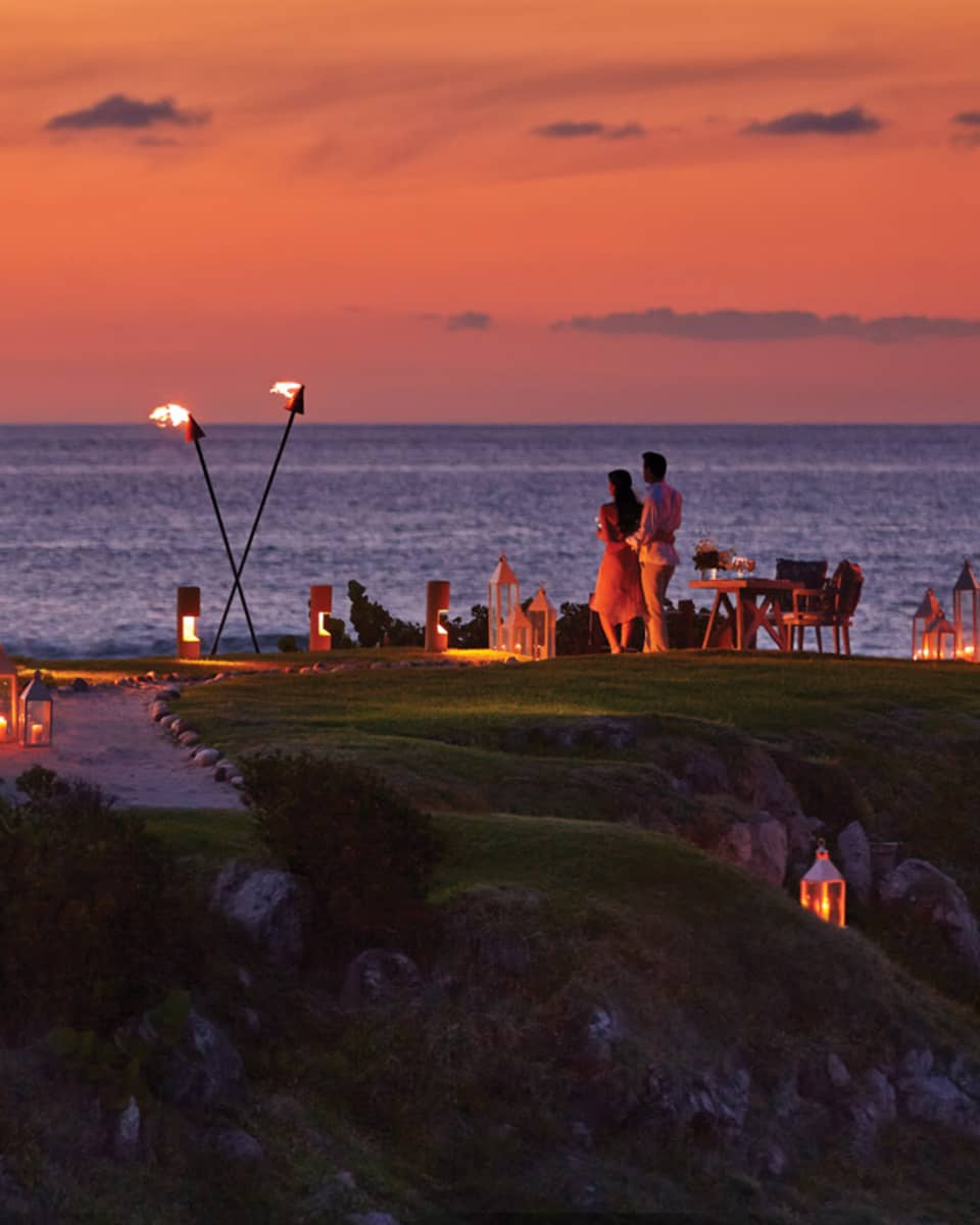 A couple stands on The Rock outpost at dusk, overlooking the ocean, with private dining table with lanterns and torches 