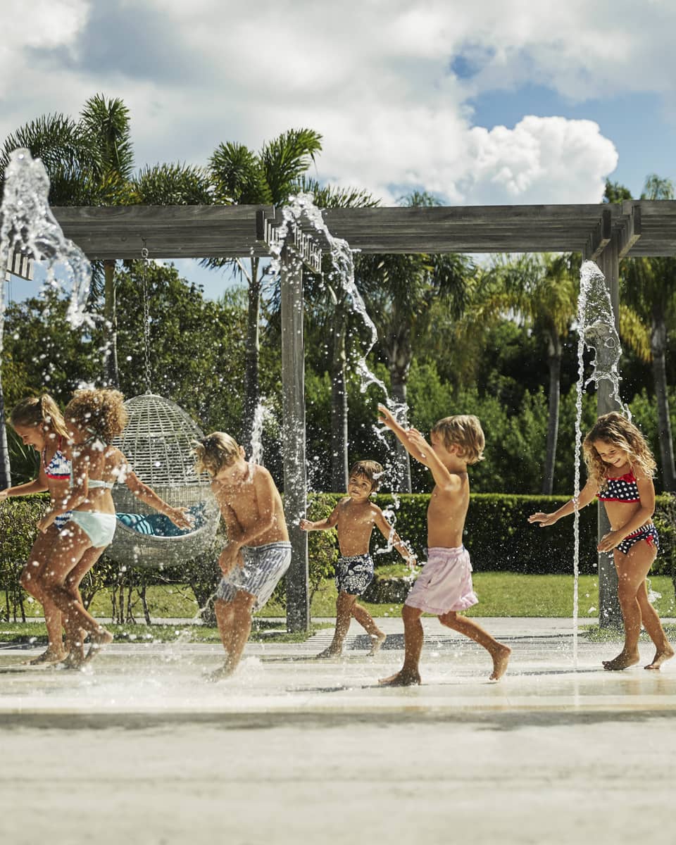 Kids in swimsuits laugh and run under outdoor splash pad, fountain