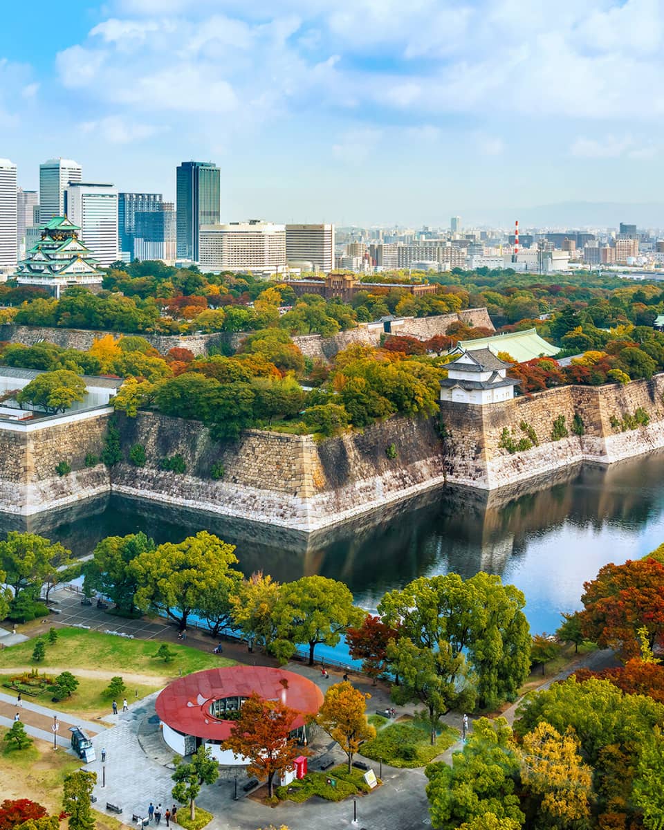 Osaka Castle rises amid lush autumn-coloured trees within a stone fortress surrounded by a moat and modern high-rises.