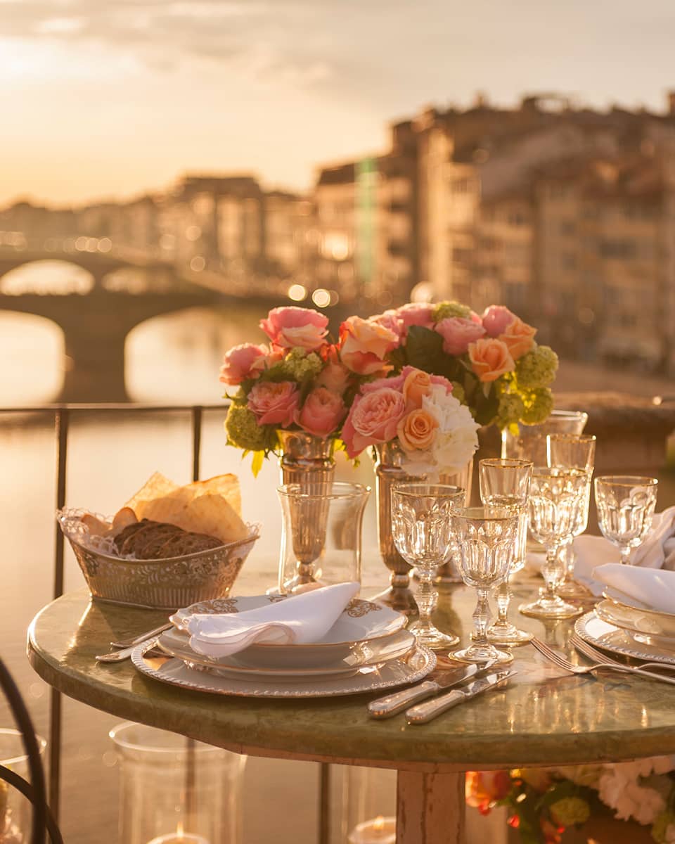 A romantic table for two is set with white porcelain plates, crystal glasses, pink roses on a balcony overlooking a bridge and historic building in Florence