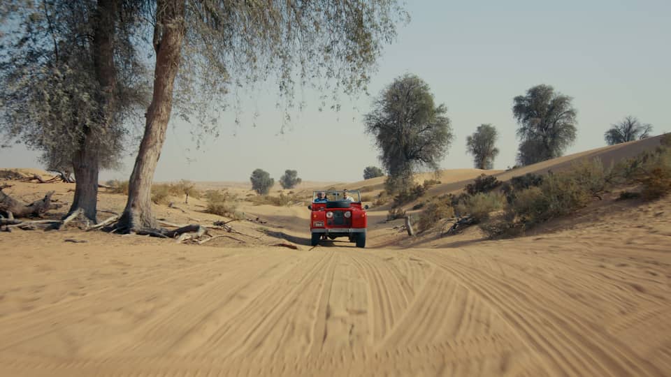 A faraway view of a red Range Rover in a dusty brown desert beneath a scrawny tree.