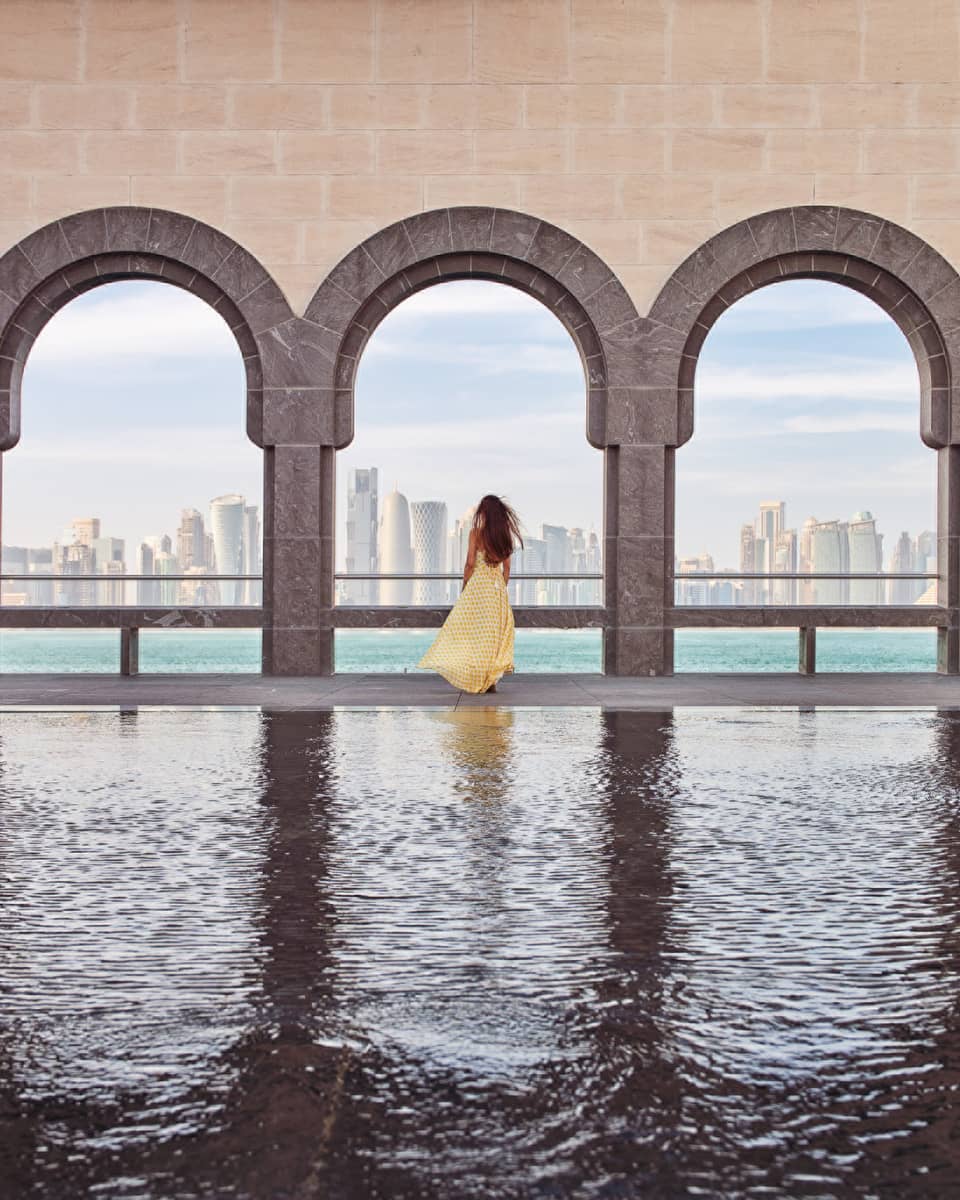 Woman wearing a long flowing yellow dress stands at the edge of a fountain beneath a wall of archways looking out onto the sea and a city skyline