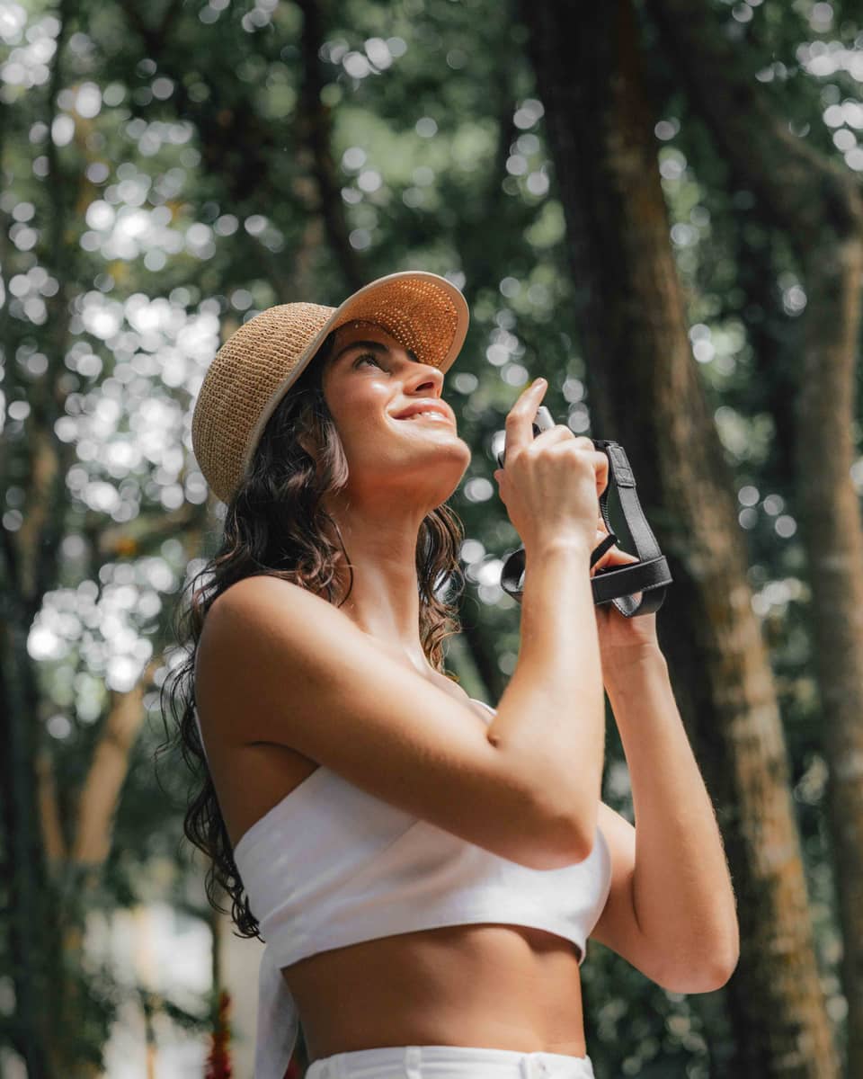 A woman stands in a tropical forest holding a camera, looking up