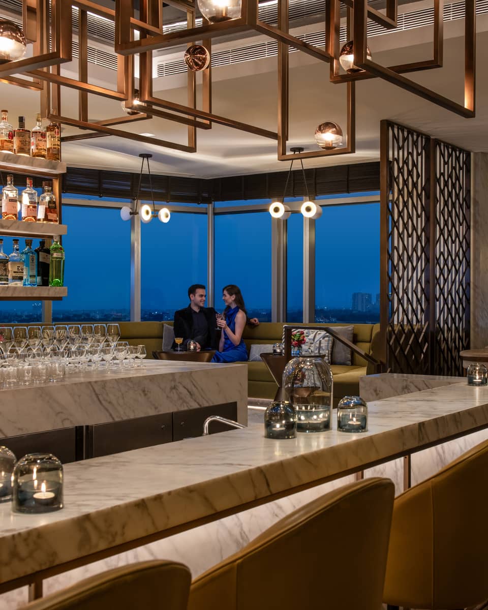 Two smiling people in formal wear sit in booth behind marble bar with liquor bottles on display