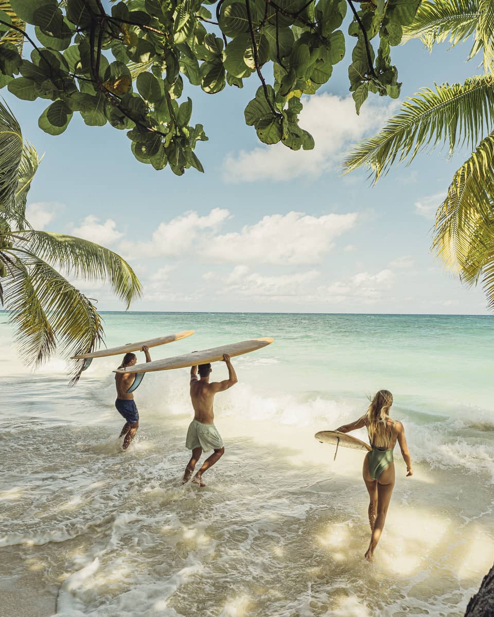 Palm trees extend over waves rushing a sandy beach, as a group of surfers with their boards bound toward the turquoise sea. 