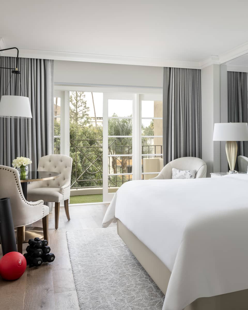 A hotel room with large open window, bed with white sheets, dresser, two chairs, an exercise ball and weights.