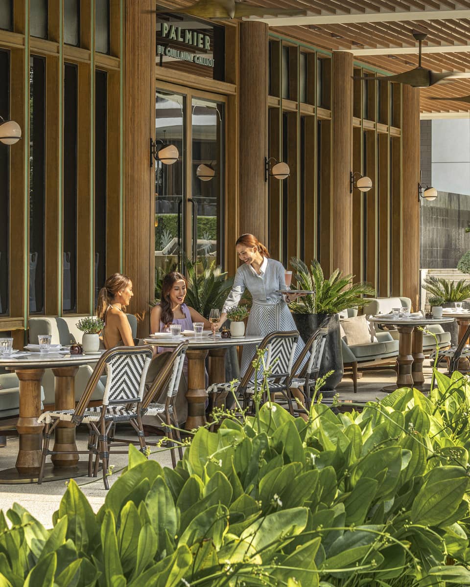 Two dining patrons are served by a waiter on the shaded patio of a restaurant