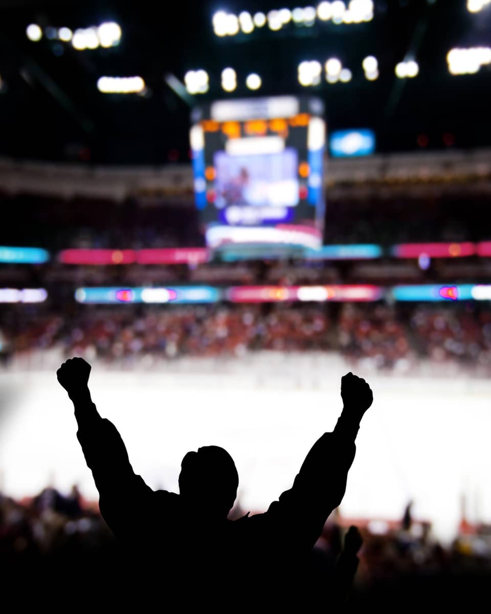 Silhouette of a fan cheering with raised arms at a hockey game in a crowded arena, with the ice rink and scoreboard in the background.
