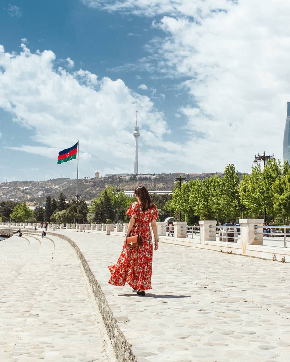 Woman wearing red dress walks along stone waterfront, skyscrapers in background