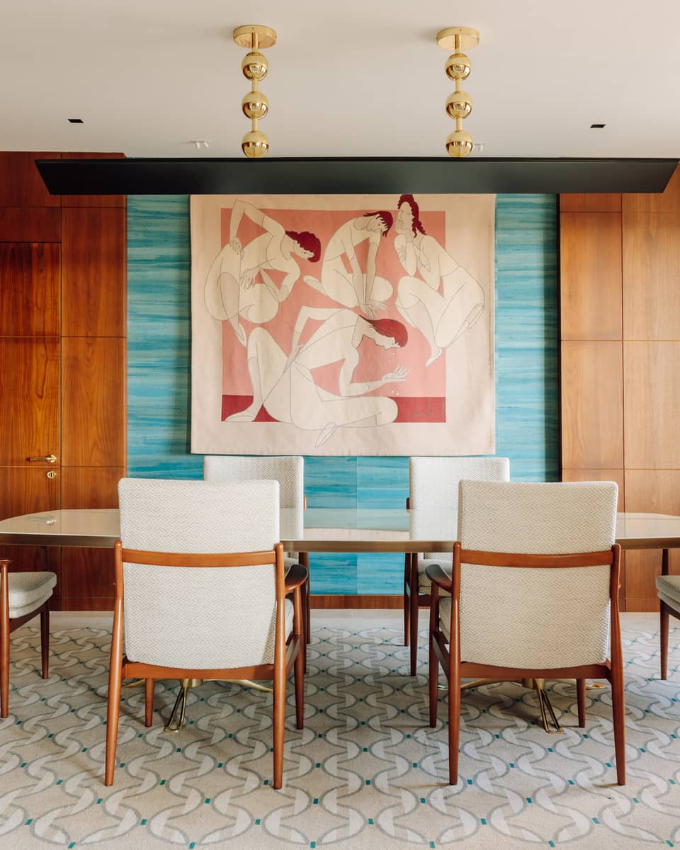 Dining room with a long table, six chairs, colorful abstract wall art and wood-paneled walls with natural light from large windows.