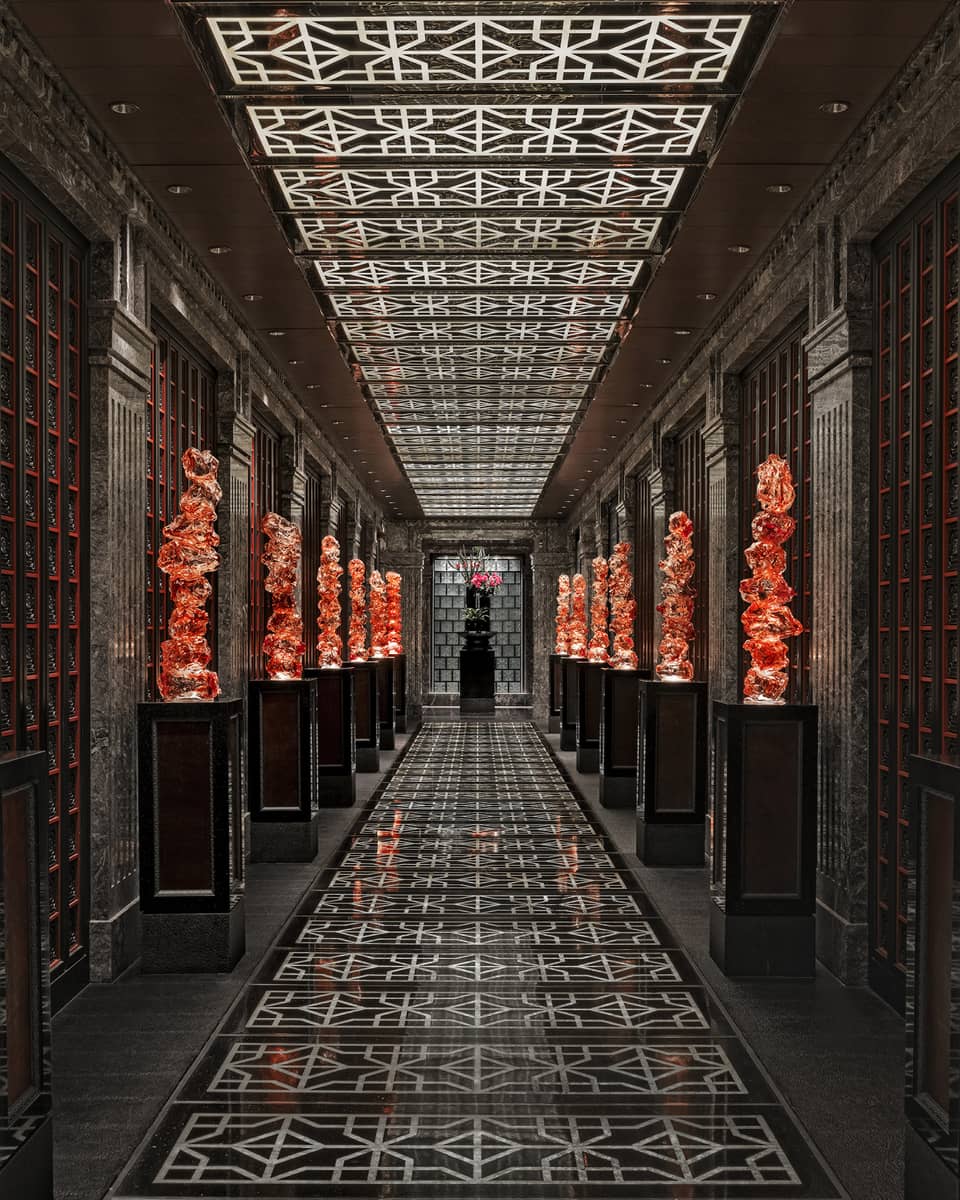 View down a long black-tiled hall lined with orange glass sculptures on pedestals, the geometric ceiling mirroring the floor.