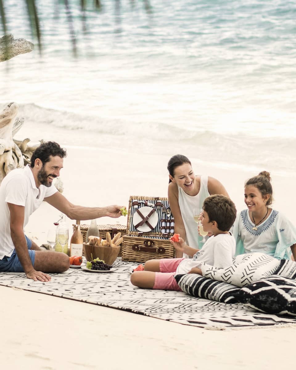 A man, woman and two children picnic on the beach just a couple feet away from the ocean. 